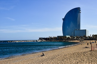 A tall, modern hotel with a distinctive curved design stands near a sandy beach. The ocean is calm, with gentle waves running parallel to the shoreline. Several people are relaxing on the beach, some walking close to the water, while a breakwater of rocks extends into the sea. Palm trees are visible near the hotel, adding to the coastal atmosphere.