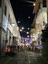 A narrow cobblestone street is illuminated by string lights and a few wall lanterns, creating a warm and inviting atmosphere. People are seated at small outdoor tables along the sides of the street, enjoying what seems to be a lively evening in a quaint town. Bicycles are parked in the foreground, adding to the charming, rustic ambiance. The buildings on either side are old-style and have simple, elegant facades. Decorations hang overhead, enhancing the festive mood.