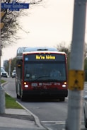 A red city bus is driving down a suburban street with a sign displaying 'We're hiring' on the front. The street is lined with trees on one side and a sidewalk on the other. A traffic sign is visible, and another vehicle follows the bus.