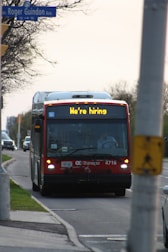 A red city bus is driving down a suburban street with a sign displaying 'We're hiring' on the front. The street is lined with trees on one side and a sidewalk on the other. A traffic sign is visible, and another vehicle follows the bus.