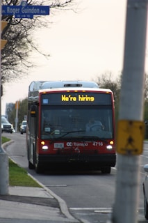 A red city bus is driving down a suburban street with a sign displaying 'We're hiring' on the front. The street is lined with trees on one side and a sidewalk on the other. A traffic sign is visible, and another vehicle follows the bus.