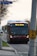 A red city bus is driving down a suburban street with a sign displaying 'We're hiring' on the front. The street is lined with trees on one side and a sidewalk on the other. A traffic sign is visible, and another vehicle follows the bus.