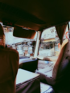 The interior of a vehicle is captured with a view through the window showing a busy street scene. Several signs with foreign language text are visible outside. The seats appear worn, suggesting use over time. The overall setting suggests an urban environment, possibly in a market area.