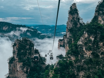 Cable cars traverse between towering sandstone pillars shrouded in mist. Lush green vegetation accents the rugged cliffs, while a vast expanse of clouds fills the valley below. The atmosphere is serene yet adventurous, inviting exploration amidst the dramatic landscape.