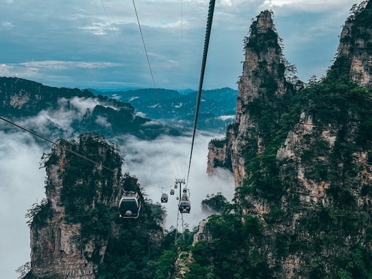 Cable cars traverse between towering sandstone pillars shrouded in mist. Lush green vegetation accents the rugged cliffs, while a vast expanse of clouds fills the valley below. The atmosphere is serene yet adventurous, inviting exploration amidst the dramatic landscape.