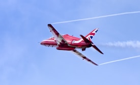 A red jet aircraft with a prominent Union Jack insignia on its tail is flying against a clear blue sky. The aircraft is emitting white smoke trails from its wingtips, creating patterns in the sky.