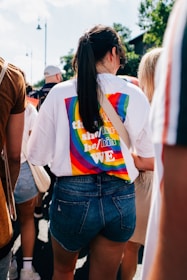 A person wearing a white shirt with a colorful rainbow pattern on the back, featuring text with various pronouns. The individual is also wearing denim shorts and has long hair tied in a ponytail. Surrounded by other people, the scene suggests a public event on a sunny day with trees and a building visible in the background.