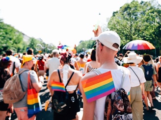 A lively outdoor gathering featuring people in colorful attire, including rainbow-patterned clothing and accessories. The scene is bright and sunny, with bubbles floating in the air and an atmosphere of celebration. Green trees line the background, and some participants carry vibrant rainbow umbrellas.