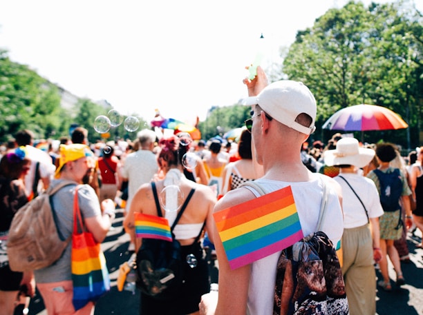 A lively outdoor gathering featuring people in colorful attire, including rainbow-patterned clothing and accessories. The scene is bright and sunny, with bubbles floating in the air and an atmosphere of celebration. Green trees line the background, and some participants carry vibrant rainbow umbrellas.