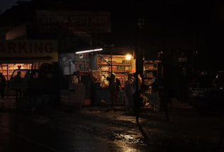 A dimly lit street shop is open at night, with shelves filled with various colorful items. A few people stand in front of the shop, illuminated by the warm glow of a single bulb. The street is wet, reflecting the light, suggesting recent rain.