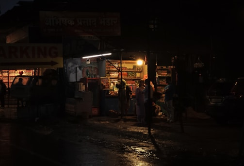 A dimly lit street shop is open at night, with shelves filled with various colorful items. A few people stand in front of the shop, illuminated by the warm glow of a single bulb. The street is wet, reflecting the light, suggesting recent rain.