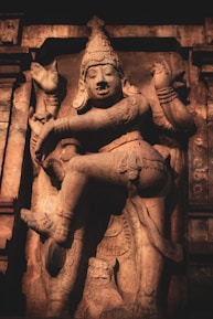 An intricately carved stone sculpture depicting a regal figure in ornate attire, striking an expressive pose. The statue features detailed accessories and a headdress, conveying a sense of grandeur and cultural significance.