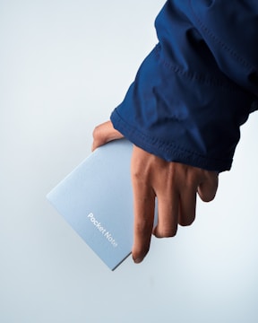 A hand holding a small, light blue notebook with the words 'Pocket Note' printed on the cover. The person is wearing a dark blue jacket, and the background is a plain, light blue color.
