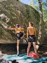 Two shirtless individuals are engaged in rock climbing, with one person actively climbing a large boulder using ropes while the other stands nearby. They are on a padded mat with climbing gear scattered around. Sunlight filters through the surrounding trees, creating a natural and adventurous outdoor setting.