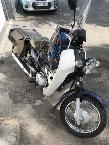 A motorcycle is parked on a street with a camouflage seat cover, a storage compartment, and a large front headlight. The vehicle's design suggests it is ready for rugged or practical usage.