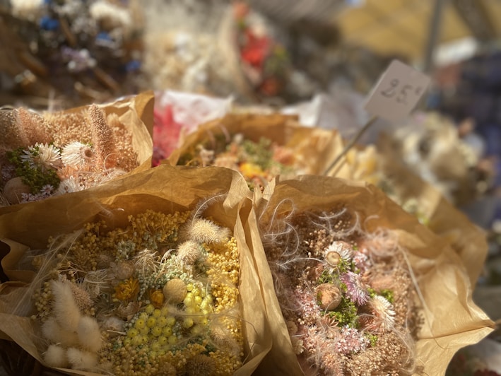 Bundles of dried flowers wrapped in brown paper, featuring a range of textures and colors such as yellows, whites, and soft pinks. The flowers include various small blooms and seed pods, structured neatly. In the background, there is a blurred sign with a price.