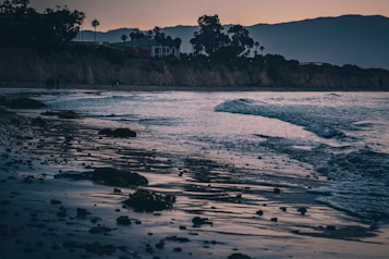 The image depicts a serene beach during sunset, with gentle waves lapping against the shore. The shoreline is scattered with rocks and seaweed, and there are silhouettes of people walking in the distance. The sky is a gradient of soft orange and purple hues, with silhouetted palm trees and cliffs in the background.