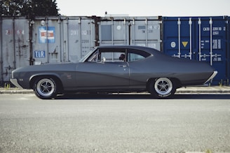 A classic gray muscle car is parked on an asphalt surface in front of stacked shipping containers. The car has a sleek, vintage design with a two-door coupe body style and prominent wheel rims. The background features industrial metal containers, some in blue and others in rusted white, with numbers and symbols marked on them.