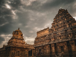 A majestic ancient temple with intricate carvings is set against a dramatic sky filled with dark, swirling clouds. The temple structures showcase detailed sculptures and architectural elements typical of historical Indian temple design.