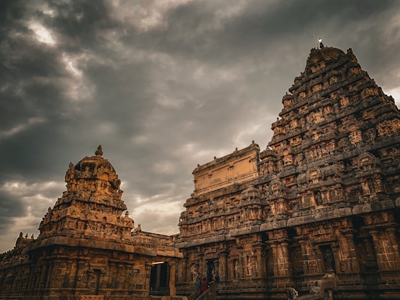 A majestic ancient temple with intricate carvings is set against a dramatic sky filled with dark, swirling clouds. The temple structures showcase detailed sculptures and architectural elements typical of historical Indian temple design.