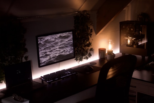 A dimly lit workspace features a computer setup with a monitor displaying a screen saver of ocean waves. The desk is adorned with subtle greenery, creating an organic feel. A warm light bulb glows gently, housed in a glass fixture near a desktop computer with a visible interior. A mechanical keyboard and a mouse are also present on the desk.