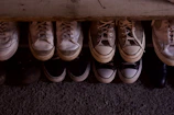 A collection of various sneakers and shoes is neatly arranged in two rows on a textured surface. The shoes vary in colors, predominantly featuring shades of white, beige, and black, with visible signs of wear indicating they have been used frequently.