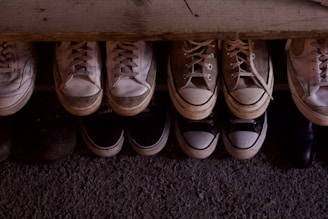 A collection of various sneakers and shoes is neatly arranged in two rows on a textured surface. The shoes vary in colors, predominantly featuring shades of white, beige, and black, with visible signs of wear indicating they have been used frequently.