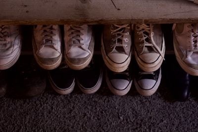A collection of various sneakers and shoes is neatly arranged in two rows on a textured surface. The shoes vary in colors, predominantly featuring shades of white, beige, and black, with visible signs of wear indicating they have been used frequently.