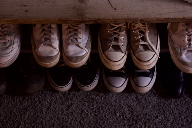 A collection of various sneakers and shoes is neatly arranged in two rows on a textured surface. The shoes vary in colors, predominantly featuring shades of white, beige, and black, with visible signs of wear indicating they have been used frequently.