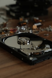 An opened hard drive lies on a wooden surface, displaying its internal components, including the reflective disk platter and the actuator arm assembly. In the background, there are various other dismantled electronic components scattered about, creating a scene of disassembly or repair.