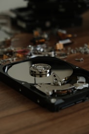 An opened hard drive lies on a wooden surface, displaying its internal components, including the reflective disk platter and the actuator arm assembly. In the background, there are various other dismantled electronic components scattered about, creating a scene of disassembly or repair.