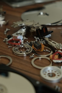 Various electronic components and metallic parts are scattered on a wooden surface, including circuit board pieces, screws, and circular metal rings.