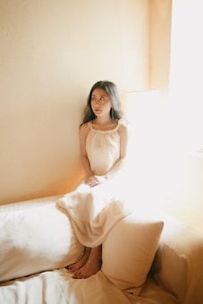 A person is sitting on a white couch in a softly lit room. They are wearing a long, light-colored dress and have long dark hair. The sunlight coming through a window creates a warm, peaceful atmosphere.
