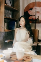 A woman in a white dress is seated on a wooden chair in a cozy room with rustic decor. Shelves on the wall hold books and a piece of art featuring a lotus flower is visible behind her. The foreground includes a wooden table with teacups and other items, suggesting a peaceful setting.
