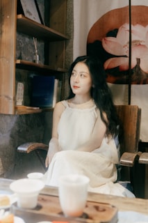 A woman in a white dress is seated on a wooden chair in a cozy room with rustic decor. Shelves on the wall hold books and a piece of art featuring a lotus flower is visible behind her. The foreground includes a wooden table with teacups and other items, suggesting a peaceful setting.