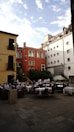An outdoor restaurant with several tables covered in white tablecloths is situated in a charming courtyard surrounded by colorful buildings. The architecture includes a mix of red, yellow, and white facades with ornate windows. Several waiters in white uniforms and a few diners are present. There are green plants and trees adding a touch of nature.