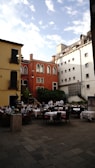 An outdoor restaurant with several tables covered in white tablecloths is situated in a charming courtyard surrounded by colorful buildings. The architecture includes a mix of red, yellow, and white facades with ornate windows. Several waiters in white uniforms and a few diners are present. There are green plants and trees adding a touch of nature.