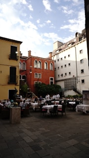 An outdoor restaurant with several tables covered in white tablecloths is situated in a charming courtyard surrounded by colorful buildings. The architecture includes a mix of red, yellow, and white facades with ornate windows. Several waiters in white uniforms and a few diners are present. There are green plants and trees adding a touch of nature.