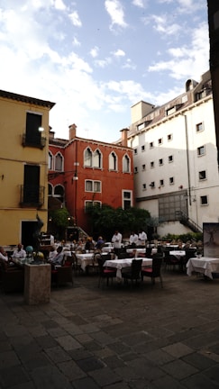 An outdoor restaurant with several tables covered in white tablecloths is situated in a charming courtyard surrounded by colorful buildings. The architecture includes a mix of red, yellow, and white facades with ornate windows. Several waiters in white uniforms and a few diners are present. There are green plants and trees adding a touch of nature.
