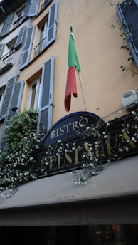 A rustic building facade with green shutters decorated with flowering vines. An Italian flag hangs prominently above a restaurant sign labeled 'Bistro'.