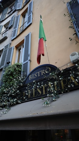 A rustic building facade with green shutters decorated with flowering vines. An Italian flag hangs prominently above a restaurant sign labeled 'Bistro'.