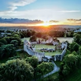 An aerial view of an ancient castle with fortified walls surrounded by lush greenery. The castle courtyard contains some outdoor furniture and tents. Beyond the castle, a town stretches out into the horizon, with a scenic sunset casting an orange glow over the landscape. The sky features scattered clouds, adding depth to the scene.