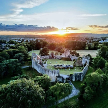 An aerial view of an ancient castle with fortified walls surrounded by lush greenery. The castle courtyard contains some outdoor furniture and tents. Beyond the castle, a town stretches out into the horizon, with a scenic sunset casting an orange glow over the landscape. The sky features scattered clouds, adding depth to the scene.