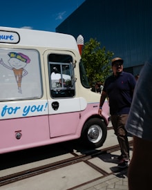 An ice cream truck with colors white and pink is parked outdoors. On the side, there is a large sticker of an ice cream cone character wearing a hat labeled 'Mr. Whippy.' A man in a dark shirt and brown pants wearing a cap is walking beside the truck. The sky is clear and blue, and there are a few trees and a modern building in the background.