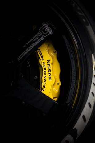 A close-up view of a yellow Nissan branded brake caliper with Brembo carbon ceramic labeling on a car wheel. The surrounding area includes parts of the black rim and tire with some branding visible. The lighting highlights the vibrant yellow of the brake caliper against the darker shades of the wheel.