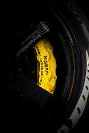 A close-up view of a yellow Nissan branded brake caliper with Brembo carbon ceramic labeling on a car wheel. The surrounding area includes parts of the black rim and tire with some branding visible. The lighting highlights the vibrant yellow of the brake caliper against the darker shades of the wheel.