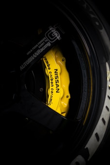 A close-up view of a yellow Nissan branded brake caliper with Brembo carbon ceramic labeling on a car wheel. The surrounding area includes parts of the black rim and tire with some branding visible. The lighting highlights the vibrant yellow of the brake caliper against the darker shades of the wheel.