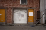 A brick building facade featuring a garage door with a mural of palm trees and the text 'HAVE A NICE DAY.' The garage door is flanked by two yellow doors, each with the street name 'PARK ROW AVE' painted vertically. One of the yellow doors has a white rectangular patch over part of it. Above the garage door, there is a small window.