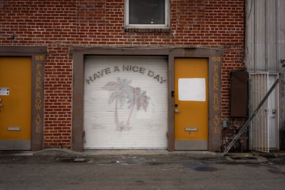 A brick building facade featuring a garage door with a mural of palm trees and the text 'HAVE A NICE DAY.' The garage door is flanked by two yellow doors, each with the street name 'PARK ROW AVE' painted vertically. One of the yellow doors has a white rectangular patch over part of it. Above the garage door, there is a small window.