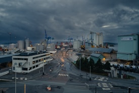 A large industrial port area with numerous storage tanks, cranes, and large buildings spread across the landscape. The sky is overcast with dark, moody clouds, and the ground appears wet, suggesting recent rainfall. Blue cranes are prominently visible, and there are several roads and pathways leading into the area. The atmosphere seems calm with no visible people or vehicles.
